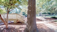 This expansive outdoor common area is defined by its mature, towering trees that provide generous canopy coverage across the grounds, creating a shaded and natural atmosphere throughout the space. The landscape is layered with pine straw mulch and leaf coverage beneath the trees, contributing to its established, park-like character. A wooden deck with a light-painted railing and staircase serves as a functional transition point within the grounds, offering elevated access and a structured gathering area amid the natural surroundings. Picnic tables are positioned beneath the tree canopy, providing dedicated outdoor seating areas ideal for recreational use. A notable stone retaining wall runs along one section of the property, adding a textured, natural architectural element that helps define the terrain. The grounds include additional landscaping features such as ornamental shrubs, decorative boulders, and multi-stem trees that introduce visual variety. Surrounding residential buildings are visible in the background, constructed in brick and featuring their own balconies. The overall ambiance is calm and verdant, with dappled sunlight filtering through the tree canopy and open grassy areas complementing the more densely wooded sections of the space.