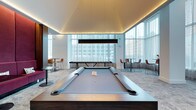 This contemporary amenity space is designed with both style and leisure in mind. At its center sits a full-size billiards table with a sleek gray felt surface, finished in a modern two-tone design that serves as the room's focal point. Suspended above it, a minimalist rectangular pendant light provides focused illumination, complementing the warm cove lighting that traces the perimeter of the tray ceiling, adding depth and a refined ambiance to the space.

Floor-to-ceiling windows line the far wall, flooding the room with abundant natural light while framing impressive views of the surrounding urban skyline. Sheer white curtains soften the light without obstructing the cityscape. The neutral-toned carpet grounds the space and contributes to its understated elegance.

Flanking the billiards table are bistro-style high-top tables paired with modern cognac leather bar stools, offering comfortable spots for socializing or casual seating. A shuffleboard table positioned near the windows adds another layer of entertainment. The overall design is clean, bright, and cohesive, striking a balance between a polished aesthetic and a relaxed, social atmosphere.