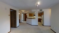 This single-floor residence offers a practical and well-organized open-concept layout that connects the living area and kitchen seamlessly. The kitchen is a standout feature, equipped with warm honey-toned wood cabinetry, a built-in microwave, a stainless steel refrigerator, and a range. A curved peninsula island serves as a natural divider between the kitchen and the living space, providing additional workspace and casual seating potential. Track lighting mounted on the ceiling highlights the kitchen area, while flush-mount fixtures illuminate the adjoining hallway. The flooring throughout is light-toned carpeting, lending the space a soft, comfortable feel. White walls and a textured ceiling complement the wood accents, creating a neutral and inviting ambiance. A hallway extends from the main living area, leading to additional rooms and providing glimpses of doors that suggest bedroom and bathroom access. The overall layout is efficient and thoughtfully designed, with clearly defined zones for cooking, dining, and living. The space presents a clean, move-in-ready environment suited for everyday functionality.
