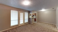 This single-floor residential unit offers a practical and comfortable layout designed for everyday living. The living room serves as the heart of the space, featuring warm neutral-toned carpet, a classic brick fireplace with a white mantle, and a glass-paneled exterior door flanked by windows that invite natural light into the room. A pass-through opening connects the living area to the kitchen, enhancing the open feel between the two spaces.

The galley-style kitchen is efficiently arranged with warm wood cabinetry lining both walls, light-toned countertops, a double-basin stainless steel sink, and white appliances including a range and refrigerator. Tile flooring and a window at the end of the kitchen add brightness to the functional workspace. A peninsula counter with upper cabinets serves as a visual divider between the kitchen and the adjacent dining or secondary living area.

The bedroom is a well-proportioned, carpeted room with a large window providing natural light and ample wall space for furnishings. A textured popcorn ceiling is consistent throughout the unit.

The bathroom features a split-style layout with a vanity and mirror on one side and a separate water closet housing a full tub and shower combination, toilet, and window. Tile flooring completes the practical design of this well-organized space.