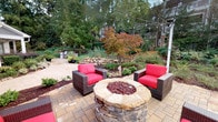 This single-story residence seamlessly blends comfortable interior living with thoughtfully designed outdoor spaces. The exterior presents a classic grey-sided architectural style with white trim accents, pitched rooflines, and manicured landscaping featuring colorful seasonal plantings, ground lighting, and stone column details. A nearby pool area adds to the community amenities visible from the grounds.

The outdoor patio serves as a standout feature, centered around a substantial stone-crafted fire pit with decorative red glass accents. Two bold red-cushioned lounge chairs frame the fire pit, all set atop a patterned paver surface. Overhead string lighting and mature trees, including a striking Japanese maple, create an inviting atmosphere. Tiered stone garden beds filled with diverse plantings complete the serene outdoor setting.

Inside, the living area features wood-style flooring, neutral grey walls with crisp crown molding, and warm ambient lighting. The space accommodates a plush sofa, accent chairs, and a patterned area rug, with bold artwork adding visual interest. The open-concept layout flows into a dining area with pendant lighting and a kitchen pass-through featuring granite countertops. Additional built-in cabinetry with granite surfaces provides functional storage throughout the interior.