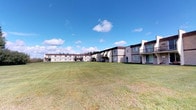 This single-story residence is part of a multi-building complex that features a mix of architectural styles, including low-rise and two-story structures with neutral-toned exteriors. The buildings are arranged to frame a generously sized, well-maintained open lawn that stretches across the grounds, providing a sense of openness and space rarely found in densely developed communities. The grassy common area offers an inviting outdoor environment with plenty of room for recreation or leisurely enjoyment under open skies.

The exterior of the complex presents a practical, straightforward design with features such as covered entryways, balconies on upper-level units, and large windows that allow natural light to filter into the living spaces. The single-floor layout of this particular unit offers convenient, step-free access, making it an accessible and easy-to-navigate living space.

The surrounding grounds benefit from a calm, open atmosphere, with views of the expansive lawn and the wider sky visible from multiple vantage points. The setting suggests a quiet, community-oriented environment with ample outdoor space. Interior images were not available, but the single-floor design typically supports a comfortable, functional layout suited to everyday living.