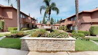 Summer Meadows Apartments presents a thoughtfully designed residential community with warm Mediterranean-inspired architecture. The exterior is characterized by earthy terracotta-hued stucco buildings accented with rich brown trim and sloped rooflines that lend the community a cohesive, welcoming aesthetic.

At the heart of the community, a beautifully landscaped courtyard serves as a central gathering point, featuring a decorative stone-raised planter overflowing with vibrant flowering plants and a classic ornamental fountain. Towering palm trees and mature evergreens frame the space, providing shade and a resort-like ambiance. Neatly trimmed hedgerows and well-maintained garden beds line the walking paths throughout the grounds, contributing to an overall sense of meticulous upkeep.

The leasing center entrance is framed by an elegant white pergola supported by classical columns, offering a shaded seating area with a cushioned bench — an inviting first impression for prospective residents. A branded entry mat reading "Summer Meadows Apartments" welcomes visitors through the glass-panel doors into the leasing office. Additional covered walkways with matching white columned pergolas extend along the perimeter, enhancing both the functionality and aesthetic of the outdoor spaces. A community directory is conveniently positioned near the main pathway, helping residents and visitors navigate the grounds with ease.