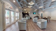 Located at 3500 Pinckney Marsh Lane in Summerville, South Carolina, this single-story community clubhouse offers a refined and well-appointed gathering space designed for both relaxation and recreation. The facility is defined by its striking coffered ceilings, which span multiple rooms and lend an architectural elegance that sets the tone throughout the entire space.

The grand lounge serves as the heart of the clubhouse, featuring soaring ceilings with a decorative chandelier, large arched windows and French doors that flood the room with natural light, and sophisticated seating arrangements centered around a glass-top coffee table. The warm hardwood flooring carries consistently through each area, unifying the design.

Adjacent to the lounge is an open social area anchored by a curved, granite-topped bar with barstool seating. Behind it, a full kitchen outfitted with dark cabinetry, stainless steel appliances, and ample counter space supports community gatherings and events. A wall-mounted television and comfortable lounge seating make this area ideal for casual socializing.

The game room provides dedicated entertainment space, complete with a handsome full-size billiards table, a round dining table, built-in bench seating, and its own wall-mounted television — all beneath the same distinctive coffered ceiling. A dedicated business center rounds out the amenities, offering dual computer workstations with granite surfaces, dark cabinetry, and a shared printer, providing a quiet and functional workspace for residents.
