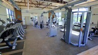 This fitness center offers a spacious, single-floor workout environment designed to accommodate a full range of exercise needs. The facility is anchored by a generous selection of professional-grade strength training machines, including multiple cable and pulley systems from a commercial equipment line, arranged to allow easy circulation and safe use throughout the floor. A dedicated free weight area lines one side of the room, with a tiered dumbbell rack providing an organized and accessible range of weights for users of all fitness levels.

The architectural design is a standout feature, with exposed wooden roof trusses soaring overhead, lending the space an open, airy character. Large windows along the perimeter flood the interior with natural light and offer views of the surrounding greenery, creating an energizing atmosphere. Ceiling fans are mounted above to maintain comfortable airflow during workouts.

The flooring consists of durable, low-pile carpet tile suited for a high-traffic fitness environment. Full-length mirrors are installed along the walls, enhancing the sense of space while allowing users to monitor their form. The overall ambiance is clean, bright, and functional, reflecting a well-maintained facility built to support a serious fitness experience.