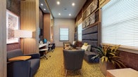 This single-floor cyber lounge is thoughtfully designed to serve as both a functional business center and a relaxed social gathering space. The room makes a strong architectural statement with soaring ceilings, tall wood-paneled columns, and a dramatic textured feature wall finished in a shimmering metallic mosaic that anchors the business center area. A wall-mounted flat-screen television and a built-in desk with multiple computer workstations and ergonomic chairs make this an ideal workspace.

The lounge area is equally well-appointed, featuring a large upholstered banquette with tall padded paneling rising to the ceiling, complemented by tufted accent chairs and an assortment of decorative pillows in neutral and olive tones. Cylindrical natural wood side tables and stump-style occasional tables add organic warmth to the contemporary palette. Intricate white sculptural wall art and framed artwork add further visual interest.

Underfoot, a bold patterned carpet in golden and amber tones unifies the space. Multiple windows dressed with roman shades and horizontal blinds allow ample natural light, while recessed ceiling lighting and pendant fixtures provide a warm, layered ambiance. Lush tropical plants are placed throughout, enhancing the inviting atmosphere.