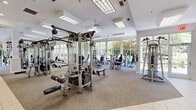 Located at 3770 George Busbee Pkwy NW in Kennesaw, Georgia, this expansive single-floor fitness center offers a comprehensive and well-appointed exercise facility. The space is organized into distinct training zones, each thoughtfully equipped to support a full range of workout routines. The cardio area houses rows of treadmills, ellipticals, and Cybex recumbent machines, all positioned alongside large windows that frame views of the surrounding greenery, creating an energizing workout environment. The strength training zone features an extensive array of cable machines, multi-station weight systems, and selectorized equipment, complemented by full-length mirrors that visually expand the space. A dedicated free weights area provides dumbbell racks, benches, medicine balls, and kettlebells, complete with rubberized flooring for safety and durability. A functional training zone with green turf flooring offers open space for dynamic movement, stretching, and conditioning exercises, with wall-mounted TVs for added entertainment. Throughout the facility, bright recessed and drop-panel lighting, crown molding, ceiling fans, and clean white walls with neutral carpeting create a polished, welcoming atmosphere. Floor-to-ceiling windows allow abundant natural light to flood each section, enhancing the overall sense of openness and space.