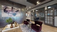 This single-floor community space presents a thoughtfully designed interior that blends modern sophistication with warm, inviting elements. The entry area welcomes visitors with light wood-look flooring, neutral grey walls, dark crown molding, and natural light filtering through slatted blinds and draped windows.

The main lounge serves as the centerpiece of the space, anchored by a dramatic large-format metallic wall panel displaying a bold piece of abstract artwork. Plush burgundy velvet accent chairs, a rustic wood coffee table, and a soft geometric area rug create an intimate seating arrangement. Recessed lighting and wall sconces contribute to a refined ambiance.

Adjacent to the lounge, a private conference or business room is accessible through glass-paned French doors. The room features exposed stacked stone accent walls, dark bar-height seating, and pendant lighting, offering a more focused, professional setting.

The fully equipped kitchen showcases rich dark-stained wood cabinetry, granite countertops, and stainless steel appliances including a range, microwave, refrigerator, and dishwasher. Wood-look flooring carries seamlessly throughout, and ample counter space makes this kitchen both functional and visually appealing.

Overall, the space combines industrial-inspired textures with warm furnishings and curated artwork, creating an atmosphere that is both polished and approachable.