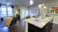 This single-floor residence offers a thoughtfully designed open-concept layout that seamlessly connects the kitchen, dining, and living areas into one cohesive and functional space. The kitchen serves as a central hub, anchored by a generous granite-topped island with bar seating, a stainless steel sink, and stylish pendant lighting overhead. Track lighting along the ceiling further enhances the kitchen's bright, welcoming atmosphere.

Rich, dark hardwood-style flooring runs throughout the main living areas, adding warmth and contrast to the light-toned walls. The dining area accommodates a full table with seating and is positioned near a set of French doors that allow abundant natural light to pour in while providing direct access to an outdoor area. Art pieces adorning the walls add a curated, finished touch to the space.

The living area is spacious and comfortable, featuring room for full-sized seating and accent furniture. A cozy area rug defines the lounge zone, while a floor lamp and sideboard contribute to the space's inviting ambiance. Overall, the residence strikes a balance between modern finishes and functional design, making it a comfortable and stylish everyday living environment.