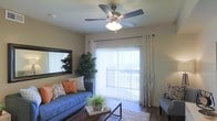 This single-floor residence offers a thoughtfully designed living space that balances comfort and functionality. The living room serves as the heart of the home, drawing in abundant natural light through a large sliding glass door fitted with sheer white curtains and vertical blinds, which open to an outdoor balcony — ideal for enjoying fresh air and outdoor views. A ceiling fan with a built-in light fixture is centrally mounted overhead, ensuring year-round comfort and efficient air circulation. The room features warm hardwood-style flooring that anchors the space with a sense of warmth and durability. A large, framed mirror on the wall enhances the sense of openness, while lush indoor plants add a refreshing, natural touch to the interior. The living area is further complemented by neutral wall tones that create a versatile backdrop for a range of décor styles. Overall, the space presents a bright, airy ambiance with practical design elements that make it both livable and inviting, suited for everyday relaxation and casual entertaining alike.
