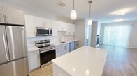 Located at 83 Pleasant Valley St in Methuen, MA, this thoughtfully designed unit offers three bedrooms and two full bathrooms across a single-floor layout. The open-concept kitchen and living area serves as the heart of the home, where warm wood-tone flooring flows throughout. The kitchen is equipped with stainless steel appliances including a French door refrigerator, range, microwave, and dishwasher, complemented by white shaker cabinetry, a subway tile backsplash, light stone countertops, a center island, and pendant lighting. The adjacent living room features a sleek fireplace with a clean white surround and sliding glass doors that invite natural light. The three bedrooms are finished with plush carpeting and windows fitted with blinds. The primary bedroom offers a comfortable footprint with access to the hallway and closet. Both bathrooms are finished with white subway tile, with one featuring a soaking tub and shower combination and the other a glass-enclosed walk-in shower with built-in shelving. Neutral tones, clean lines, and modern hardware throughout give the space a cohesive, contemporary feel.