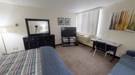 This single-floor unit offers a thoughtfully arranged layout that maximizes functionality within a compact footprint. The main living area serves as both a bedroom and lounge space, featuring wall-to-wall carpeting that adds warmth underfoot. A full-size bed with a dark wood headboard anchors the sleeping area, complemented by a matching dresser with a large mirror and a multi-drawer nightstand. A flat-screen television on a glass media stand, along with a dedicated workspace featuring a desk and chairs, ensures both entertainment and productivity needs are met. A wall-mounted HVAC unit below a wide window with vertical blinds provides climate control while allowing natural light to filter into the room. A comfortable sofa with accent pillows rounds out the living area, offering additional seating or lounging space. The unit includes a walk-in closet with built-in shelving, providing generous storage. The bathroom is neatly appointed with a toilet and a shower/tub combination concealed behind a decorative floral curtain, paired with tasteful framed wall art. Hardwood-style flooring in the entryway corridor connects the spaces seamlessly, contributing to a clean and cohesive aesthetic throughout.
