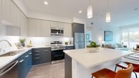 This well-appointed 2-bedroom, 2-bathroom residence showcases a clean, contemporary design across a single floor. The open-concept kitchen and living area serves as the heart of the home, featuring a striking two-tone cabinet scheme with light upper cabinets and bold dark lower cabinets, crisp white quartz countertops, a spacious island with seating, and a full suite of stainless steel appliances. Cylindrical pendant lights add a modern finishing touch overhead, while wood-look flooring flows seamlessly throughout the space. The living room is bright and inviting, with large windows framing outdoor views and filling the space with natural light. Both bedrooms are comfortably sized, with the primary bedroom offering a sliding-door closet and large windows that bring in additional daylight. The bathrooms feature sleek, dark vanities with white countertops and wood-look flooring, maintaining the home's consistent modern aesthetic. A dedicated hallway connects the bedrooms and bathrooms, providing a practical and well-organized layout. Throughout the residence, white walls and recessed lighting create a clean, airy ambiance that complements the overall contemporary style.