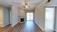 This single-floor residence offers a thoughtfully designed living space that balances comfort and style. The living room serves as the centerpiece of the home, showcasing warm hardwood-style plank flooring that runs throughout and lends a sense of continuity and elegance to the space. A classically styled fireplace with a clean white mantel serves as a natural focal point, adding both character and coziness to the room. Crown molding frames the ceiling with refined architectural detail, while a tray ceiling adds depth and dimension overhead. A ceiling fan with integrated lighting provides both comfort and functionality year-round. Natural light fills the room through a large sliding glass door, which provides direct access to an outdoor area and invites the outside in. The neutral, warm-toned walls create a versatile backdrop that complements a variety of interior styles. The open layout allows for flexible furniture arrangements, making the living area feel spacious and welcoming. The overall ambiance of the space is calm and inviting, with architectural details that elevate its everyday livability while maintaining a clean, move-in-ready aesthetic.