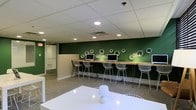 This single-floor space is designed as a functional and visually engaging shared workspace or business center. The room's most striking feature is a deep forest-green accent wall that serves as a bold focal point, adorned with white hexagonal wall-mounted shelving units arranged in an asymmetric, decorative pattern that adds both style and dimension.

Along the green wall runs a continuous wooden counter-height workstation equipped with multiple desktop computers, making it well-suited for individual focused work or collaborative tasks. Sleek white wire-frame bar stools are positioned at each station, combining an industrial aesthetic with a modern sensibility.

The flooring consists of neutral-toned carpet tiles that complement the contemporary color palette, while the drop-tile ceiling features recessed lighting that provides even, well-distributed illumination throughout the space. A white communal table occupies the foreground, offering additional flexible workspace.

The layout balances open space with defined work zones, creating an environment that feels both organized and inviting. A glass-paneled door visible at the far end provides connectivity to adjacent areas, enhancing the overall flow of the space.