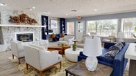 Located at 7212 Carlton Arms Dr in New Port Richey, FL, this single-story community clubhouse offers a thoughtfully designed gathering space with a welcoming, contemporary aesthetic. The entrance greets visitors with a staffed reception desk, light wood-look flooring, and vibrant framed artwork adorning two-toned blue and white walls throughout.

The expansive main lounge is anchored by a dramatic floor-to-ceiling natural stone fireplace, complemented by a built-in navy blue bookshelf and a dedicated workstation area with seating for residents. Comfortable lounge furniture in neutral tones is artfully arranged around a mix of round coffee tables, creating an inviting atmosphere for socializing or relaxing.

Large windows along multiple walls flood the interior with natural light and provide views of the outdoor surroundings, including a fenced pool area. A sun-drenched seating nook lines the windowed wall, offering additional casual seating with mid-century modern accent chairs.

The clubhouse also features a well-appointed kitchenette with a long quartz-topped bar counter, navy cabinetry, a stainless steel commercial ice maker, and a sink — ideal for community events. A separate dining area with round tables and upholstered chairs provides a dedicated space for gatherings. A quiet waiting area with classic wood chairs and curated artwork rounds out this versatile community facility.