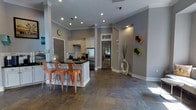 This well-appointed single-floor space showcases a thoughtfully designed open-concept layout that seamlessly connects the kitchen and living areas. The kitchen is a standout feature, boasting white shaker-style cabinetry, subway tile backsplash, dark granite countertops, and stainless steel appliances, including a full-size refrigerator. A central kitchen island with a dark countertop provides generous prep and dining space, accompanied by copper-finished metal bar stools that add a warm industrial touch. The flooring throughout is a rich, multi-toned natural stone tile, lending the space an earthy yet sophisticated character. Walls are painted in a cool grey tone, complemented by crisp white crown molding and baseboards that frame each room with architectural precision. Recessed lighting illuminates the interior, while a glass-paned door at the rear allows natural light to filter into the space. Decorative wall-mounted geometric shelving adds a modern artistic element, while sculptural accents on the floor contribute to the space's curated aesthetic. Structural columns further define the open layout while maintaining the airy, connected feel of the floor plan.