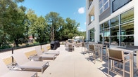 This outdoor amenity space is a thoughtfully designed terrace that offers a versatile setting for both relaxation and social gatherings. The terrace is finished with a smooth, light-toned concrete surface that provides a clean, contemporary aesthetic throughout the area.

The space is organized into distinct functional zones. A lounge area features modern sling-style chaise lounges with sleek aluminum frames, positioned to take full advantage of the open-air environment. Adjacent to this, a dining zone offers high-top bar tables paired with matching sling chairs, ideal for casual outdoor meals or social mingling. A gas grill station is also available, making the terrace well-suited for outdoor cooking and entertaining.

The terrace runs alongside a modern multi-story building characterized by a crisp white exterior and large floor-to-ceiling glass windows and doors, which allow natural light to flow between the indoor and outdoor spaces. Upper-level balconies are visible above, reinforcing the building's contemporary architectural language.

Mature trees line the perimeter of the property, providing a natural backdrop and a degree of greenery and privacy. Under open skies and bathed in natural sunlight, the terrace offers a welcoming outdoor retreat that balances modern design with comfortable, practical amenities.