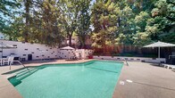 Located at 7747 Greenback Ln in Citrus Heights, California, this outdoor recreational area offers a well-appointed communal space designed for relaxation and leisure. At the heart of the space is a generously sized swimming pool with clear turquoise water, complemented by an adjacent hot tub for additional relaxation. The pool deck is finished with a textured aggregate surface and is lined with multiple lounge chairs and patio umbrellas, providing ample space for sunbathing and outdoor socializing. A dedicated BBQ station with a full-sized gas grill is built into the perimeter wall, alongside outdoor dining furniture shaded by a striped market umbrella — ideal for al fresco gatherings. The surrounding enclosure features a painted block wall with decorative stone accents, adding a structured yet welcoming aesthetic to the environment. Mature trees frame the entire area, offering natural shade and a lush, private atmosphere. The backdrop reveals a multi-story residential building with solar panels and private balconies overlooking the pool area. Wooden fencing and landscaped greenery further enhance the sense of seclusion. Overall, this outdoor space delivers a resort-like ambiance within a well-maintained residential community setting.
