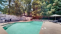 Located at 7747 Greenback Ln in Citrus Heights, California, this outdoor recreational area offers a well-appointed communal space designed for relaxation and leisure. At the heart of the space is a generously sized swimming pool with clear turquoise water, complemented by an adjacent hot tub for additional relaxation. The pool deck is finished with a textured aggregate surface and is lined with multiple lounge chairs and patio umbrellas, providing ample space for sunbathing and outdoor socializing. A dedicated BBQ station with a full-sized gas grill is built into the perimeter wall, alongside outdoor dining furniture shaded by a striped market umbrella — ideal for al fresco gatherings. The surrounding enclosure features a painted block wall with decorative stone accents, adding a structured yet welcoming aesthetic to the environment. Mature trees frame the entire area, offering natural shade and a lush, private atmosphere. The backdrop reveals a multi-story residential building with solar panels and private balconies overlooking the pool area. Wooden fencing and landscaped greenery further enhance the sense of seclusion. Overall, this outdoor space delivers a resort-like ambiance within a well-maintained residential community setting.