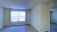 This single-floor space presents a straightforward and functional layout. The main living area is finished with wall-to-wall carpeting in a neutral gray-blue tone, providing a soft underfoot surface that extends throughout the room. The walls are painted in a clean, off-white shade, contributing to a bright and open atmosphere. A large window fitted with vertical blinds serves as the primary source of natural light, allowing diffused daylight to illuminate the interior. The textured popcorn ceiling adds a subtle visual element overhead. Electrical outlets are conveniently positioned along the baseboards, and white baseboard trim cleanly defines the transition between the walls and floor. The room is currently unfurnished, making it easy to assess the available space and envision a variety of furniture arrangements. Overall, the space offers a clean, neutral canvas suitable for a range of uses and décor styles.