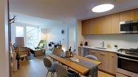 This thoughtfully designed single-floor residence showcases a clean, modern aesthetic throughout its open-plan layout. The kitchen is outfitted with warm wood cabinetry, stainless steel appliances, and a sleek countertop with an integrated sink, offering both functionality and style. Flush cabinet hardware and crisp white walls keep the look streamlined and contemporary.

The dining area centers around a natural wood table accompanied by a mix of modern chairs, creating an inviting space for gathering. A textured table runner and curated tableware add warmth and personality to the setting.

Flowing seamlessly from the dining space, the living area benefits from generous natural light streaming through a large window, illuminating the room and offering views of the surrounding greenery. A statement lounge chair in vibrant green anchors the sitting area alongside a low-profile coffee table, lending the space a relaxed yet design-forward character.

Consistent light wood flooring runs throughout, unifying each zone of the open floor plan. Overhead, modern flush and pendant lighting provide a warm, ambient glow. Thoughtful decorative touches — including a sunburst wall clock and carefully chosen furnishings — contribute to a cohesive, well-appointed interior that balances comfort with contemporary design.