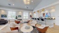 Located at 7027 Park Mill Dr in Plain City, Ohio, this single-story clubhouse offers a sophisticated and welcoming environment designed for both comfort and social engagement. The open-concept layout is anchored by herringbone-patterned hardwood flooring that flows seamlessly throughout the space, lending a refined, contemporary aesthetic.

The main lounge area is bathed in natural light from oversized windows and features soaring vaulted ceilings adorned with a statement starburst chandelier. Stylish swivel chairs and curated seating arrangements invite casual conversation, while vibrant artwork adds color and personality to the crisp white walls.

A standout fireplace serves as a dramatic focal point in a secondary lounge, framed by a striking floor-to-ceiling agate stone panel above a dark stacked-stone surround. Tufted leather sofas and a low oval coffee table complete this inviting retreat.

A well-appointed wet bar runs along one side of the main lounge, featuring a geometric-patterned base, black countertops, bar seating, and gold pendant lighting overhead. An additional service area provides white cabinetry, black marble countertops, a beverage refrigerator, and a mounted display screen.

A quieter nook near the entrance offers a classic leather lounge chair and ottoman beside a tall indoor tree, creating a cozy, private reading corner. The overall ambiance is polished, modern, and thoughtfully designed for community gathering.