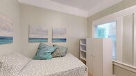 Located at 1010 10th St in Sacramento, CA, this compact studio unit is thoughtfully designed to maximize every square foot of its single-room footprint. The space is organized into distinct functional zones, making efficient use of its cozy layout. Warm-toned hardwood flooring runs throughout, lending a clean and cohesive feel to the interior. The sleeping area accommodates a twin bed with under-bed storage, complemented by a freestanding white wardrobe unit with open shelving — providing practical storage without overwhelming the space. Coastal-themed wall art adds a light, calming aesthetic to the soft sage-green walls. The kitchenette area is well-appointed with light gray shaker-style cabinetry, open shelving, a stainless steel sink with countertop space, a full-size stainless steel refrigerator, and a microwave — offering essential cooking amenities in a compact footprint. A full-length mirror and a sleek black accent chair round out the furnishings. Natural light enters through multiple windows fitted with blinds, brightening the space considerably. Crown molding details add a subtle architectural character. A wall-mounted climate control unit ensures year-round comfort. This efficiently designed space is well-suited for a single occupant prioritizing location and functionality in the heart of Sacramento.