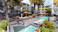 This dynamic outdoor complex is centered around a thoughtfully designed pool area that serves as the social heart of the property. Two distinct pool areas are visible across the grounds — one featuring a spacious, geometric swimming pool framed by decorative floral patterns on the surrounding deck, set within a courtyard enclosed by multi-story buildings with glass-railed balconies and lush mature trees. The second pool area takes on a more resort-inspired character, with a tiered layout that incorporates a spa or hot tub, a wooden deck platform, and multiple shaded cabanas furnished with curtains, lounge seating, and umbrella tables. Tall palm trees and layered landscaping add a Southern California ambiance throughout. A row of sleek white chaise lounges lines the pool's edge, offering a relaxed sunbathing setting. Adjacent to the pool area is a semi-enclosed recreational space featuring a full ping-pong table beneath a woven pendant light. A vivid, large-scale mural in the style of Keith Haring — depicting colorful dancing figures on a bold yellow background — serves as a striking focal point, infusing the area with an energetic and artistic character. The overall atmosphere is playful, modern, and resort-like.