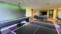 This single-floor entertainment space is purpose-built for recreation and social engagement. The room is anchored by a full-size ping pong table positioned at the center, complemented by a sleek shuffleboard table alongside it, offering a variety of game options for guests and visitors. Recessed ceiling lighting illuminates the space evenly, while accent lighting in vibrant hues — including green and purple tones — adds a lively, energetic ambiance to the environment. The walls feature a mix of neutral and bold tones, contributing to the room's modern and dynamic aesthetic. Toward the back of the space, a large flat-screen television is mounted above a low-profile media console, creating a dedicated viewing and lounge area that extends the entertainment possibilities beyond active gaming. The flooring is finished in a dark tone, tying the room's design together with a cohesive, contemporary feel. The layout is thoughtfully organized to allow multiple activities to take place simultaneously without overcrowding, making this space well-suited for group events, casual hangouts, or competitive play. Overall, the space strikes a balance between function and atmosphere, delivering a well-equipped recreational environment.