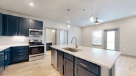 This single-floor unit showcases a thoughtfully designed interior with a cohesive contemporary aesthetic. The kitchen serves as the focal point, featuring striking navy blue shaker-style cabinetry that extends floor to ceiling, complemented by crisp white quartz countertops and a classic white subway tile backsplash. A generously sized central island includes an undermount sink with a brushed nickel faucet and pendant lighting above, making it ideal for both meal preparation and casual dining. Stainless steel appliances, including a French door refrigerator, electric range, over-the-range microwave, and dishwasher, complete the culinary space. Light wood-look flooring flows throughout the main living areas, adding warmth and continuity. The bedrooms offer plush carpet underfoot, ceiling fans, and large windows that bring in natural light and outdoor views. One room features direct exterior access through a glass-panel door, adding functional versatility. The bathrooms mirror the kitchen's design language, featuring navy cabinetry, white quartz vanity tops, brushed nickel fixtures, glass-enclosed walk-in showers, and light hardwood-style flooring, creating a clean and polished finish throughout.
