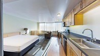This single-floor studio unit offers a thoughtfully arranged open-plan layout that seamlessly integrates the sleeping, living, and kitchen areas into one cohesive space. Large bay-style windows span the far wall, flooding the interior with natural light and creating a bright, airy atmosphere throughout the day. The sleeping area is positioned to take advantage of this natural light, accompanied by a bedside dresser for convenient storage. A cozy seating area with an upholstered armchair provides a relaxed living space just steps from the bed. The kitchen runs along the right side of the unit and is well-equipped with light wood cabinetry, dark countertops, a stainless steel sink, an electric range with an overhead microwave, and a dishwasher — offering full cooking functionality within a compact footprint. Warm hardwood-style flooring extends across the main area, complemented by a area rug that helps define the sleeping zone. The walls are finished in a soft, neutral tone, contributing to the clean and calm ambiance of the space. Flush-mount ceiling lights provide consistent illumination throughout. Overall, this studio presents a practical and comfortable living environment with efficient use of space and a modern, understated aesthetic.