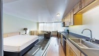 This single-floor studio unit offers a thoughtfully arranged open-plan layout that seamlessly integrates the sleeping, living, and kitchen areas into one cohesive space. Large bay-style windows span the far wall, flooding the interior with natural light and creating a bright, airy atmosphere throughout the day. The sleeping area is positioned to take advantage of this natural light, accompanied by a bedside dresser for convenient storage. A cozy seating area with an upholstered armchair provides a relaxed living space just steps from the bed. The kitchen runs along the right side of the unit and is well-equipped with light wood cabinetry, dark countertops, a stainless steel sink, an electric range with an overhead microwave, and a dishwasher — offering full cooking functionality within a compact footprint. Warm hardwood-style flooring extends across the main area, complemented by a area rug that helps define the sleeping zone. The walls are finished in a soft, neutral tone, contributing to the clean and calm ambiance of the space. Flush-mount ceiling lights provide consistent illumination throughout. Overall, this studio presents a practical and comfortable living environment with efficient use of space and a modern, understated aesthetic.