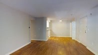 This single-floor space offers a clean and functional layout designed for comfortable everyday living. The main living area is anchored by warm-toned hardwood flooring that extends throughout, creating a cohesive and inviting atmosphere. Neutral gray and white walls provide a versatile backdrop that complements a variety of interior styles. A ceiling fan is centrally positioned in the living area, contributing both to the room's practicality and its understated aesthetic. The open floor plan allows the living space to flow naturally toward a partially enclosed kitchen area, which is defined by a low dividing wall and features white cabinetry visible in the background. This semi-open design maintains a sense of connectivity between the spaces while providing subtle separation. White baseboards frame the perimeter of the rooms, adding a crisp finishing detail. A white paneled door is visible toward the rear of the space, suggesting access to an additional room or closet. The overall ambiance is bright, clean, and move-in ready, with a straightforward layout that maximizes usable floor space across the single level.