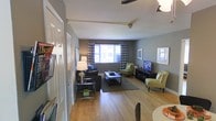 This single-floor space centers around a welcoming living area finished with warm hardwood flooring that extends throughout. The walls are painted in a cool, neutral gray tone, creating a modern and cohesive backdrop. A ceiling fan with a light fixture is mounted overhead, contributing to both comfort and ambient lighting.

The living area is generously proportioned, accommodating a full seating arrangement that includes a dark sofa, complementary green accent chairs, and a rectangular coffee table set atop a area rug. A media console along one wall houses a flat-screen television, providing a clear focal point for the room.

Natural light enters through a pair of windows dressed with bold chevron-patterned curtains, adding a decorative touch while softening the overall aesthetic. Framed artwork on the walls contributes to the curated feel of the space.

A closet with a paneled white door is visible near the entryway, offering practical storage. A dining area is partially visible in the foreground, suggesting a natural flow between living and dining spaces. Overall, the layout is functional, well-lit, and thoughtfully arranged.
