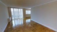 This single-floor space offers a bright and open layout characterized by classic design elements and well-maintained finishes throughout. The main living area is a standout, showcasing beautifully polished parquet hardwood flooring arranged in a distinctive block pattern that adds warmth and visual interest underfoot. Large sliding glass doors dominate one wall, dressed with full-length vertical blinds that allow for adjustable natural light and provide access to the outdoors. An additional window further enhances the natural brightness of the room, creating an airy and welcoming atmosphere. The walls are finished in a neutral tone and accented by crisp white crown molding and clean baseboards, lending the space a timeless and polished look. The open floor plan allows for flexible furniture arrangement, making the living area functional and adaptable to various uses. The overall ambiance is clean, light-filled, and well-appointed, with the gleaming parquet floors reflecting natural light to amplify the sense of space. This property combines classic architectural detailing with practical, open living — ideal for those who appreciate understated elegance and bright interiors.