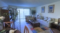 This two-floor residence offers a thoughtfully arranged living space that balances comfort and functionality. The main living area serves as the heart of the home, featuring plush, neutral-toned sofas arranged around a dark wood and glass coffee table, creating an inviting setting for relaxation or entertaining. A colorful patterned area rug anchors the seating arrangement, adding a warm, casual touch to the carpeted floor. Natural light flows generously into the room through a large sliding glass door that opens to an outdoor balcony or patio, offering views of surrounding greenery. A decorative wrought-iron room divider with leafy accents adds an artistic, ornamental element near the entryway, while a framed artwork piece on the wall and a classic table lamp contribute to the room's understated, homey character. A potted plant near the window further enhances the connection to the outdoors. Adjacent to the living area, a glimpse of a dining space is visible, suggesting a practical and open flow between living and dining areas. The textured ceiling and soft, neutral wall tones throughout lend the space a clean, cohesive aesthetic across both floors.