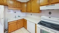 This single-floor space is organized into a functional, open layout that connects the kitchen and living areas seamlessly. The kitchen is equipped with warm oak cabinetry offering generous storage, white laminate countertops, a double sink, tile flooring with a neutral backsplash, and white appliances including a range, dishwasher, and refrigerator. A track lighting system provides focused illumination throughout.

The living area features neutral wall-to-wall carpeting, a ceiling fan with an integrated light, and a track lighting fixture, creating a versatile and well-lit environment. A large window fitted with vertical blinds allows natural light to fill the space and offers outdoor views.

The bedrooms are carpeted and each includes a ceiling fan with a light fixture, baseboard heating units, and windows with horizontal blinds that provide both natural light and privacy. The clean white walls keep the rooms feeling bright and open.

The bathroom is finished with tile flooring, a white vanity with sink, a full tub and shower combination with a tiled surround featuring decorative accent tiles, a recessed ceiling light, and a small window for natural ventilation. The overall aesthetic throughout the space is clean, neutral, and practical.