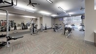 This fitness center is a comprehensively equipped facility designed to support a wide range of workout styles and fitness goals. The single-floor space is organized into distinct training zones, creating an efficient and functional layout throughout.

The cardio zone features a variety of machines including treadmills, upright and recumbent stationary bikes, a rowing machine, and a stair climber, positioned near large windows that bring in natural light and outdoor views. A bank of full-length mirrors along one wall enhances the sense of openness and allows users to monitor their form.

The strength training area is anchored by a multi-station cable machine, a selection of weight benches, and dumbbell racks, complemented by a bold red accent wall that adds energy to the space. A dedicated functional fitness zone includes a heavy punching bag, TRX suspension trainers, stability balls, kettlebells, and a rubber-floored open area ideal for dynamic movement and stretching.

Throughout, the facility features patterned carpet tiles with red accents, durable rubber flooring in high-activity zones, and bright linear overhead lighting. Ceiling fans are distributed across the space to maintain airflow and comfort. Wall-mounted televisions are positioned for visibility from key workout areas, rounding out a thoughtfully designed fitness environment.
