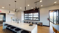 Located at 210 SW Century Dr in Bend, Oregon, this modern single-floor commercial space offers a thoughtfully designed open-concept layout ideal for collaborative and professional use. The centerpiece of the main area is a large white quartz island with an integrated sink, bar-height seating on both sides, and sleek cylindrical pendant lights suspended above — creating a natural hub for gathering and informal meetings. Warm hardwood flooring flows throughout, complementing the clean white walls and contemporary finishes. A standout architectural feature is the oversized glass-panel garage-style door that opens to reveal a bar-height ledge with a row of stools, seamlessly connecting the interior to an outdoor patio area and flooding the space with natural light and views of the surrounding landscape. Adjacent to the main area, a dedicated conference room accommodates a full-size meeting table and multiple chairs, with large windows on two walls offering abundant daylight and exterior views. A separate lounge nook features a bold navy accent wall, wall-mounted display screen, and colorful seating, providing a more intimate setting for presentations or informal gatherings. Additional high-top tables and varied seating zones throughout the space offer versatile configurations to suit a range of professional needs.