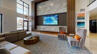 This contemporary two-floor amenity space is designed with a clean, modern aesthetic that emphasizes open layouts, polished tile flooring, and thoughtful details throughout. The grand lobby greets visitors with a striking feature wall of mixed-material cladding, a large-format video display, and plush seating arrangements set against high ceilings and abundant natural light streaming through expansive floor-to-ceiling windows. A staffed reception desk anchors the space, while an adjacent lounge area provides comfortable seating with views to the outdoors, where palm trees and a vibrant urban streetscape can be seen. The clubroom is well-equipped with a large central island, pendant lighting, a mounted flat-screen display, and a bar area finished in an elegant geometric tile backsplash. A dedicated game room offers a full-size pool table, shuffleboard, and a poker table beneath a striking wood-slat ceiling installation with Edison-bulb accent lighting. Workstation areas with built-in desks and seating provide functional co-working spots. Corridors on both floors are adorned with framed artwork, lending a gallery-like quality to the circulation spaces. Throughout, the design balances warm wood tones, neutral palettes, and contemporary furnishings to create an inviting and versatile communal environment.
