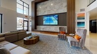 This contemporary two-floor amenity space is designed with a clean, modern aesthetic that emphasizes open layouts, polished tile flooring, and thoughtful details throughout. The grand lobby greets visitors with a striking feature wall of mixed-material cladding, a large-format video display, and plush seating arrangements set against high ceilings and abundant natural light streaming through expansive floor-to-ceiling windows. A staffed reception desk anchors the space, while an adjacent lounge area provides comfortable seating with views to the outdoors, where palm trees and a vibrant urban streetscape can be seen. The clubroom is well-equipped with a large central island, pendant lighting, a mounted flat-screen display, and a bar area finished in an elegant geometric tile backsplash. A dedicated game room offers a full-size pool table, shuffleboard, and a poker table beneath a striking wood-slat ceiling installation with Edison-bulb accent lighting. Workstation areas with built-in desks and seating provide functional co-working spots. Corridors on both floors are adorned with framed artwork, lending a gallery-like quality to the circulation spaces. Throughout, the design balances warm wood tones, neutral palettes, and contemporary furnishings to create an inviting and versatile communal environment.