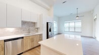 This well-appointed 2-bedroom, 2-bathroom residence is designed with a clean, modern aesthetic across a single floor. The open-concept kitchen and living area serves as the heart of the home, featuring a large white quartz island, two-tone cabinetry with warm wood lower cabinets and crisp white uppers, a subway tile backsplash, stainless steel appliances including a French door refrigerator, and recessed lighting. The living room benefits from large windows with horizontal blinds that allow generous natural light, light wood-look flooring, a ceiling fan, and a private entry door with a transom window above. A built-in desk and shelving nook adds a functional workspace near the kitchen. The hallway, finished with the same light hardwood flooring and recessed lighting, connects the main living spaces to the private quarters. The primary bedroom offers a comfortable footprint, carpet flooring, a ceiling fan, and a walk-in closet with built-in rod and shelving. The second bedroom is similarly finished with carpet, a ceiling fan, and a bright window with exterior views. Both bathrooms are well-appointed; one features a soaking tub with marble-style tile surround, wood-toned vanity, and hardwood-style flooring. Two dedicated closets provide ample storage throughout the residence.