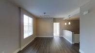 This single-floor space showcases an open-concept layout that seamlessly connects the living area to the kitchen. The living space features dark hardwood-style flooring that contrasts elegantly with the neutral beige walls and bright white baseboards and trim, creating a clean and contemporary aesthetic throughout.

Natural light flows generously into the living area through a large window fitted with white horizontal blinds, brightening the space considerably. A modern track lighting fixture is mounted on the ceiling, providing focused illumination across the main living area.

The kitchen is partially visible to the right, distinguished by a raised breakfast bar counter with a dark countertop surface and white cabinetry below. Three stylish pendant lights hang above the counter, adding a warm, inviting glow to the kitchen zone and serving as a decorative focal point.

A doorway visible toward the rear of the space leads to an additional room, suggesting functional separation between living and private areas. The overall ambiance is modern, clean, and move-in ready, with a neutral color palette that offers versatility for a variety of interior design preferences.