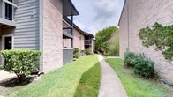 This single-floor unit is part of a well-maintained multi-story brick building complex, where lush, manicured lawns and mature trees line a concrete walkway along the exterior. The grounds offer a verdant, serene setting with balconies visible on upper levels, contributing to the community's established character.

Inside, the interior reflects a thoughtfully curated aesthetic. The dining area serves as a focal point, anchored by a striking round green marble table paired with woven rattan chairs atop a natural fiber rug. Recessed lighting illuminates the cream-toned walls, which display framed large-format artwork, adding an artistic sensibility to the space. Dark hardwood-style flooring runs throughout, grounding the warm, eclectic atmosphere.

A notable design feature is the inclusion of both a rustic, aged double door and a sleek sliding barn door on a black rail system, providing distinct spatial transitions between rooms. The overall interior palette — neutral walls, dark floors, and curated decor — creates a cohesive, gallery-like ambiance that balances sophistication with warmth. The layout flows efficiently, making thoughtful use of the single-floor footprint.