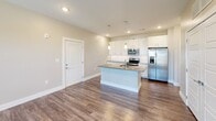 Located at 1980 Icon Way in Knoxville, Tennessee, this one-bedroom, one-bathroom unit offers a well-designed single-floor layout with modern finishes throughout. The open-concept kitchen and living area serves as the heart of the space, featuring a central island with a quartz-style countertop, white shaker cabinetry, a subway tile backsplash, stainless steel appliances including a refrigerator with ice maker, and pendant lighting overhead. Wood-look plank flooring flows seamlessly through the entire unit, complementing the neutral, warm-toned walls. A set of double panel doors in the main area provides access to additional storage or utility space. The bedroom is a comfortable, well-proportioned room with natural light from dual windows with views of the surrounding landscape, and a ceiling fan for added comfort. It connects to a walk-in closet outfitted with wire shelving and hanging rods on three walls, offering generous storage capacity. The bathroom features a wide vanity with undermount sink, illuminated mirror, and a full tub with dark tile surround. The exterior showcases stone and board-and-batten siding with landscaped rock beds, situated within a residential community with scenic surroundings.