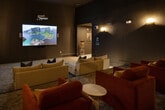 Located at 3280 Champion Ring Rd in Fort Myers, Florida, this dedicated media room delivers a fully immersive, private cinema experience within a thoughtfully designed single-floor layout. The space is anchored by a large wall-mounted projection screen positioned at one end of the room, paired with a ceiling-mounted projector that ensures a sharp, expansive viewing image. Deep charcoal and near-black wall finishes throughout create a light-controlled environment ideal for screening, while a series of warm wall sconces provide subtle, atmospheric accent lighting along the side walls. Seating is arranged in a tiered, theater-inspired layout, combining plush cream-colored sectional sofas accented with navy and blue throw pillows in the front rows, and rich rust-red upholstered armchairs in the rear. Small round side tables are interspersed throughout for added convenience. The floor is covered in a durable, patterned low-pile carpet that complements the dark, sophisticated color palette. Abstract artwork adorns one wall, adding a refined artistic touch to the cinematic setting. A connected hallway, visible from the main room, provides access to the broader facility. The overall ambiance is moody, modern, and purpose-built for entertainment.