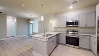 This well-appointed single-floor unit offers a thoughtfully designed open-concept layout that seamlessly connects the kitchen and living areas. The kitchen is a standout feature, showcasing granite countertops, crisp white upper cabinetry, gray lower cabinets, and a full suite of stainless steel appliances including a range, microwave, dishwasher, and refrigerator. A peninsula island with a double stainless steel sink and elegant pendant lighting adds both functionality and style. Light wood-look flooring flows throughout the main living areas, complementing the neutral gray walls and white trim. The living area is generously proportioned, featuring a ceiling fan and access to natural light through blinds-covered windows. A carpeted bedroom provides a comfortable retreat, bathed in natural light from a trio of large windows fitted with horizontal blinds. The bathroom is finished with classic white subway tile surrounding a soaking tub, gray cabinetry with granite countertops, and wood-look flooring, maintaining a cohesive and modern aesthetic. Multiple interior doors suggest ample closet and storage space. The overall design is clean, contemporary, and move-in ready, with consistent finishes and attention to detail throughout.