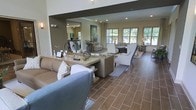 This thoughtfully designed single-floor space offers an expansive open-plan layout that seamlessly connects multiple living and lounge areas. The interior is anchored by rich tile flooring laid in a classic brick pattern, lending warmth and texture underfoot throughout the main living zones. Generous seating arrangements — including plush sofas, accent chairs, and stylishly appointed conversation areas — create distinct yet flowing spaces ideal for both relaxation and socializing.

The main living area features a large central console table adorned with curated decorative accents, framed by comfortable seating on either side. A row of wide windows along the far wall floods the space with natural light and offers picturesque views of the surrounding greenery, enhancing the open and airy atmosphere. Recessed lighting complements the natural light, ensuring the space remains bright and inviting at all times.

Beyond the main seating area, a secondary lounge zone is visible through an open architectural passageway, complete with its own casual seating and decorative artwork. Neutral tones of taupe, grey, and cream dominate the palette, accented by carefully chosen furnishings and décor that strike a balance between contemporary style and approachable comfort. The overall ambiance is refined yet welcoming, making this a versatile and elegant space.