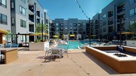 This outdoor pool area in St. Louis, MO is a well-appointed courtyard retreat designed for relaxation and social gatherings. At its center sits a generously sized swimming pool with crystal-clear water, complemented by a shallow wading shelf and multiple entry points. The surrounding deck is finished in warm-toned concrete pavers, providing ample space for sunbathing and casual gatherings, and is furnished with aluminum tables, chairs, and cushioned lounge seating throughout.

A built-in fire pit lounge anchors one corner of the courtyard, surrounded by low-profile seating walls ideal for evening socializing. An outdoor bar station with bar-height seating offers a dedicated space for entertaining. Vibrant yellow market umbrellas are positioned throughout the deck for shade, while colorful string lights are strung overhead, creating a festive ambiance that transitions seamlessly from day to evening use.

Lush greenery and potted plants are thoughtfully integrated into raised planters, adding natural softness to the contemporary hardscape. The courtyard is enclosed on multiple sides by a modern multi-story building featuring large windows, private balconies, and a clean gray-and-white façade with bold dark trim. Large glass sliding doors provide convenient indoor-outdoor access, connecting the pool deck directly to interior common spaces.
